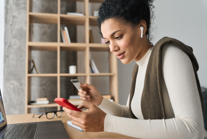 A woman sitting at a desk scrolling on her phone with a credit card in her hand.