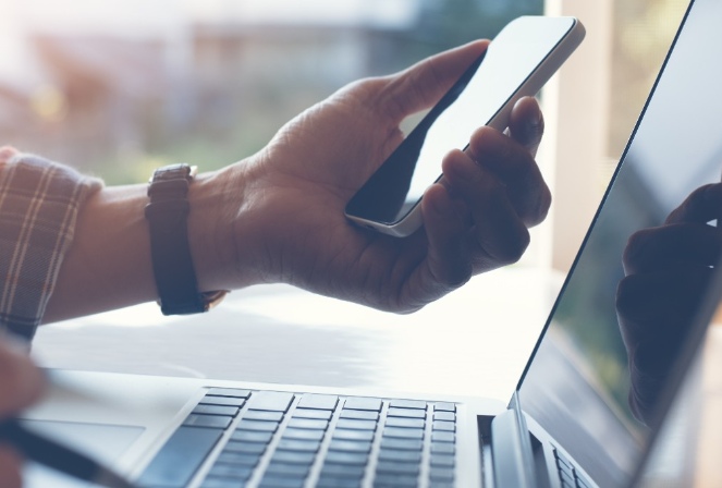 a person holding their mobile phone and working on their laptop