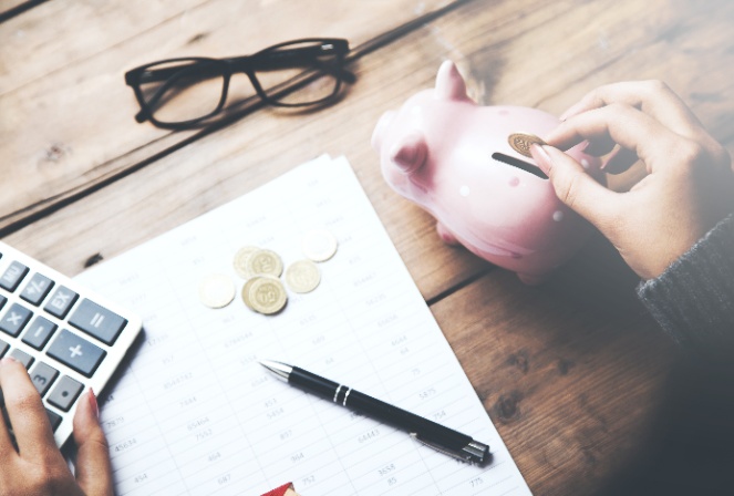 A wooden desk with a calculator, glasses, coins, a notepad and a hand putting a coin in a piggy bank.